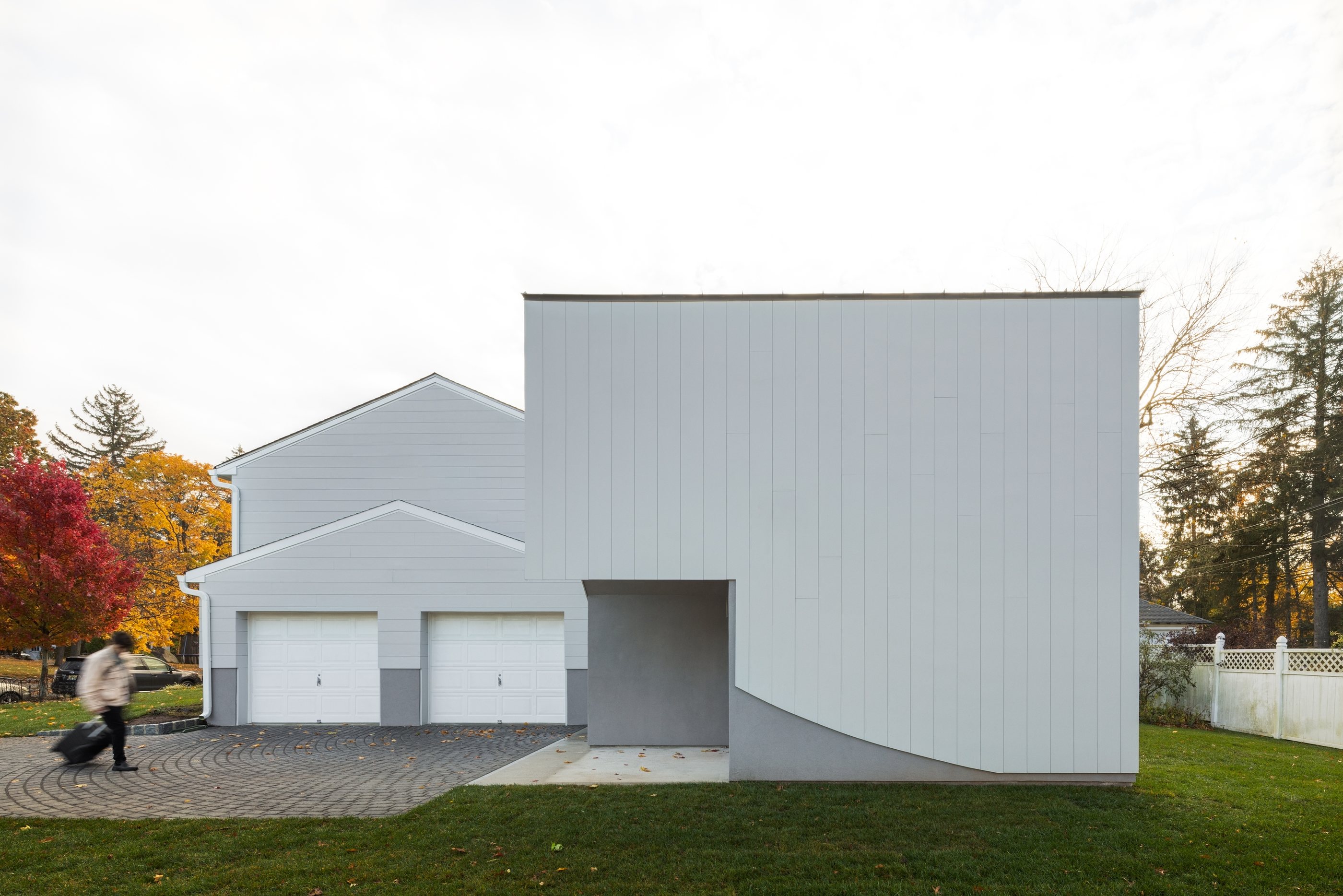 The Swoop ADU's white facade beside the existing home's two-car garage, seen from the street in autumn