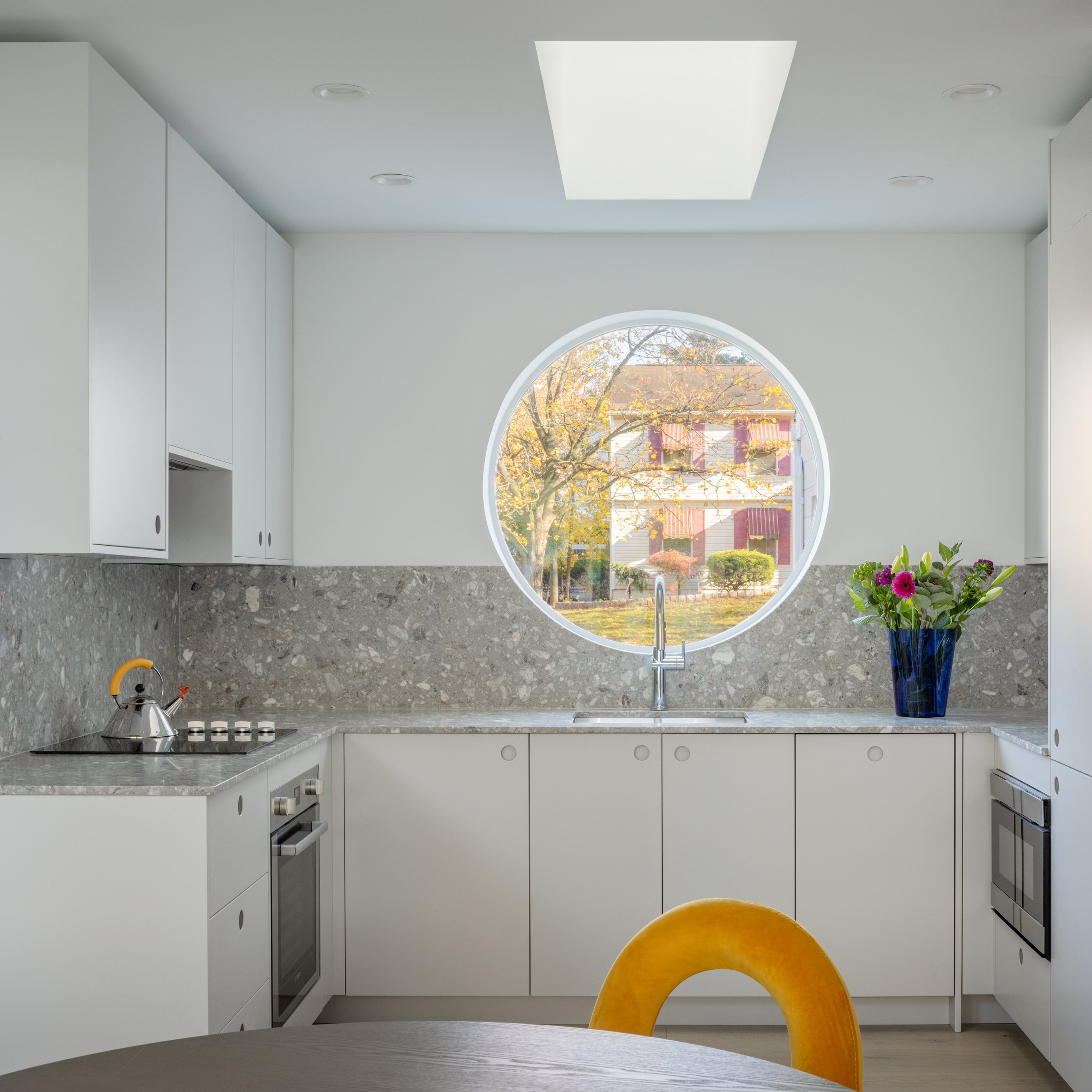 The Swoop ADU kitchen with white Reform cabinetry, gray terrazzo counters and backsplash, and a round window over the sink