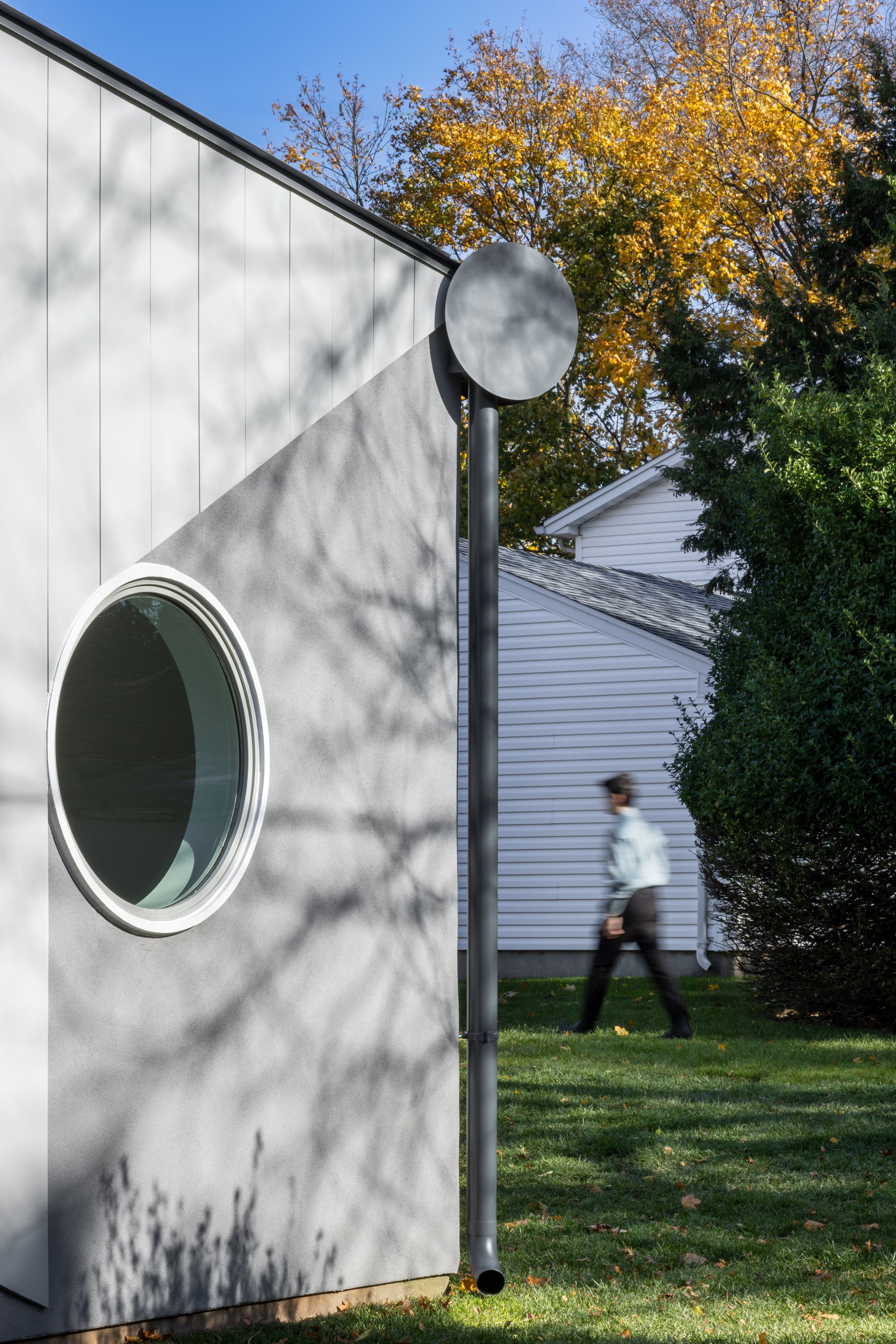 Close view of the round porthole window set into the Swoop ADU's curved white wall, with a neighbor passing on the lawn
